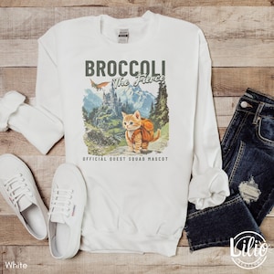 K&ouml;nnte beinhalten: Wei&szlig;er Langarm-Pullover mit einem Aufdruck eines Katers mit Rucksack und dem Text "Broccoli The Fierce" und "Official Quest Squad Mascot".