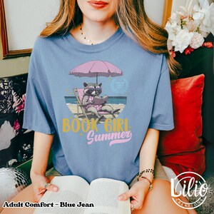 May include: A blue jean t-shirt with a graphic of a raccoon reading a book on a beach chair under a pink umbrella. The text "Book Girl Summer" is printed on the shirt.