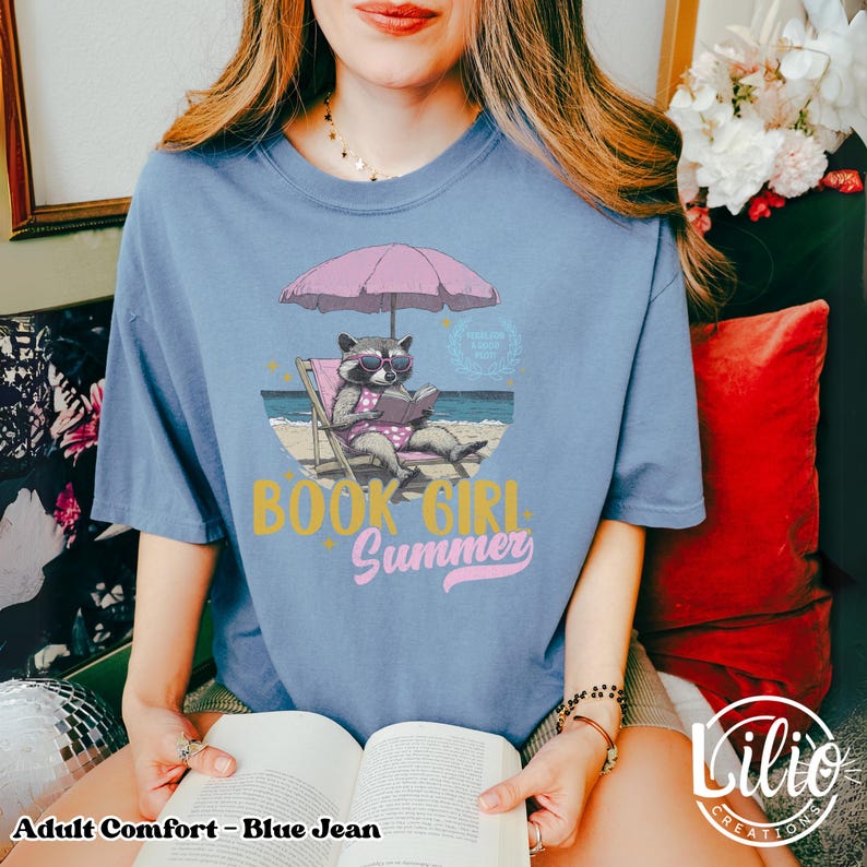 May include: A blue jean t-shirt with a graphic of a raccoon reading a book on a beach chair under a pink umbrella. The text "Book Girl Summer" is printed on the shirt.