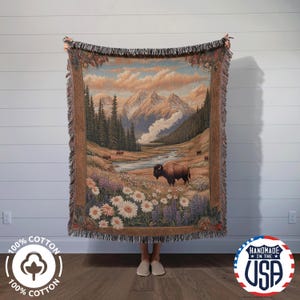 May include: A woven tapestry throw blanket features a scenic landscape with a river, mountains, and a brown bison in a field of wildflowers. The tapestry has a fringe border and is made of 100% cotton.