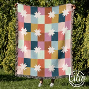 May include: A colorful woven blanket with a star pattern. The blanket features a grid of squares in various colors, including pink, blue, orange, and green, with white starburst designs. The edges have a fringed trim.
