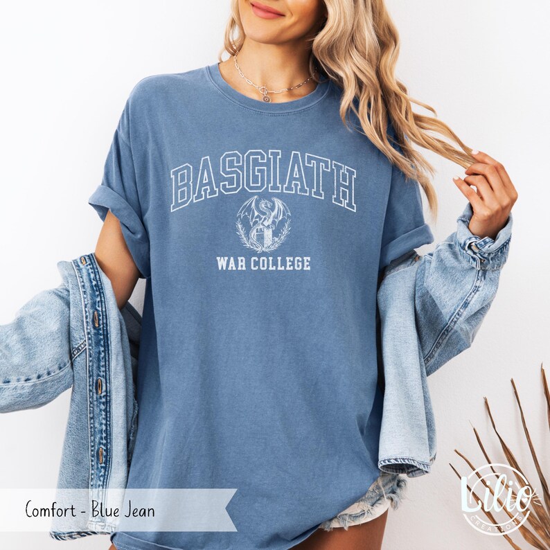 K&ouml;nnte beinhalten: Blaues Jeans-T-Shirt mit dem Wort "BASGIATH" in wei&szlig;en Blockbuchstaben. Darunter eine Grafik eines Drachen und die Worte "WAR COLLEGE". Das Shirt hat eine bequeme Passform.