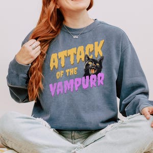 May include: A blue sweatshirt with the text "Attack of the Vampurr" in yellow and purple letters. A black cat with yellow eyes is featured in the graphic.