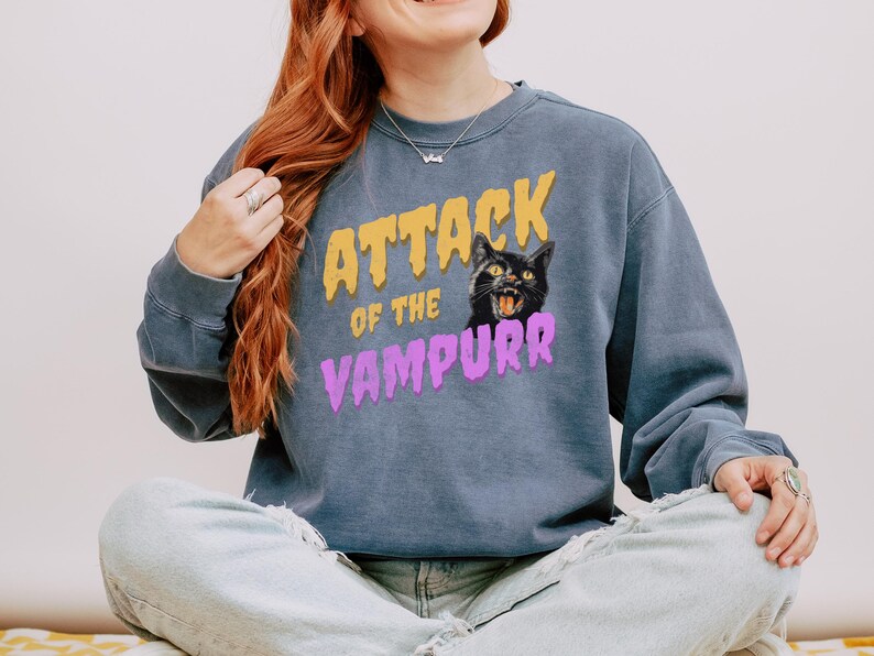 May include: A blue sweatshirt with the text "Attack of the Vampurr" in yellow and purple letters. A black cat with yellow eyes is featured in the graphic.