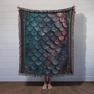 K&ouml;nnte beinhalten: Eine gewebte Wolldecke mit einem dunkelblauen und violetten Schuppenmuster, das von einem goldfarbenen Rand umrahmt wird. Die Decke hat einen Fransenrand.
