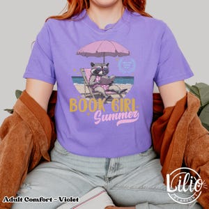 May include: A purple t-shirt with a graphic of a raccoon wearing sunglasses and reading a book under a pink umbrella on a beach. The text "Book Girl Summer" is written in gold and pink letters.
