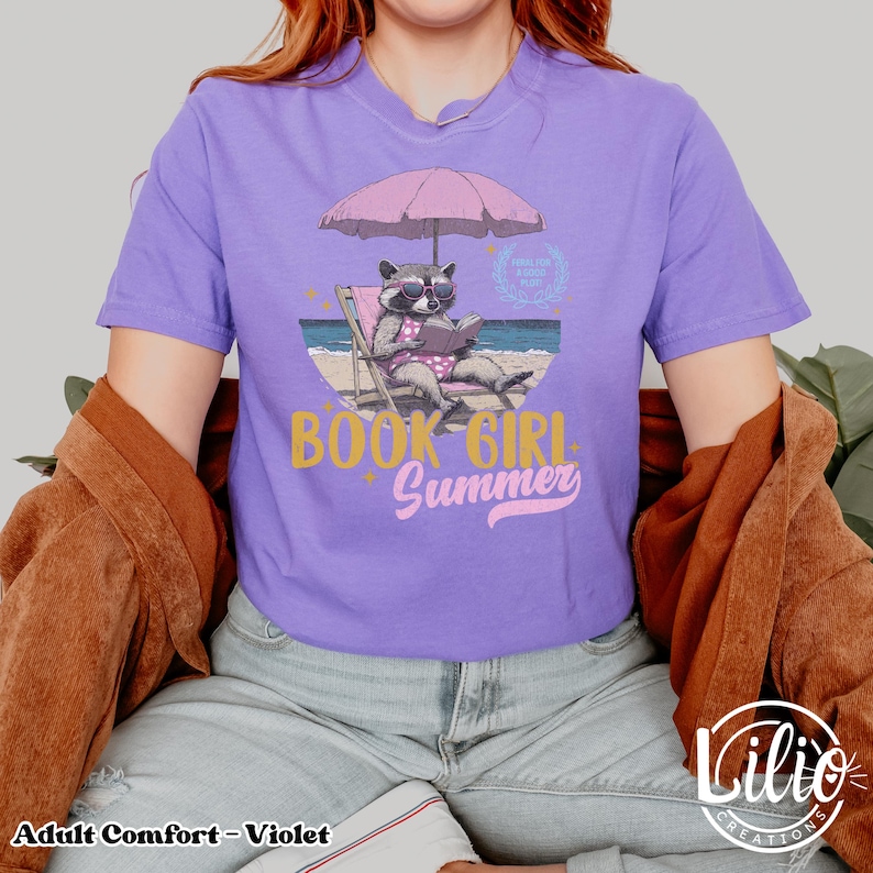 May include: A purple t-shirt with a graphic of a raccoon wearing sunglasses and reading a book under a pink umbrella on a beach. The text "Book Girl Summer" is written in gold and pink letters.