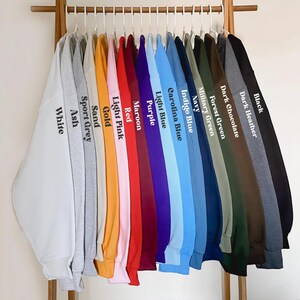 K&ouml;nnte beinhalten: Eine Reihe von 16 Sweatshirts, die an einem Holzgestell h&auml;ngen und eine Vielzahl von Farben zeigen, darunter Wei&szlig;, Asche, Sportgrau, Sand, Gold, Hellrosa, Rot, Weinrot, Lila, Hellblau, Himmelblau, Indigo, Marine, Milit&auml;rgr&uuml;n, Waldgr&uuml;n, Dunkelbraun, Dunkelgrau und Schwarz. Jedes Sweatshirt hat den Farbnamen auf dem &Auml;rmel aufgedruckt.
