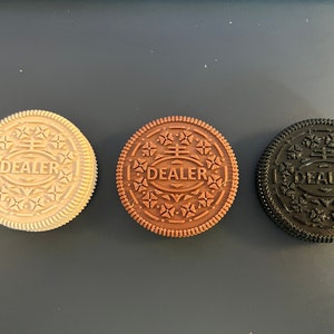 May include: Three round cookies with the word "DEALER" printed on them. The cookies are in different colors: white, brown, and black.