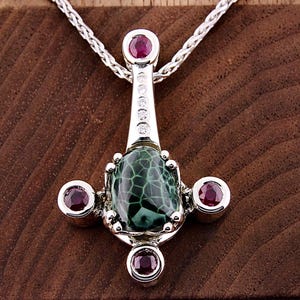 May include: A silver pendant necklace with a green and black stone, surrounded by four smaller pink stones. The pendant is attached to a silver chain.
