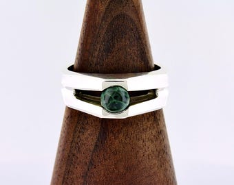 Op de afbeelding: Een zilveren ring met een geometrisch ontwerp, met een donkergroene, bolvormige steen in het midden. De ring wordt gepresenteerd op een houten ringhouder, die de moderne en minimalistische stijl laat zien. Het ontwerp van de ring is strak en eigentijds.
