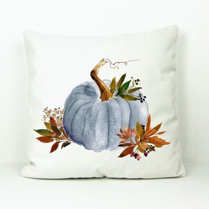 May include: A white pillow with a watercolor illustration of a blue pumpkin with brown stems and leaves. The pumpkin is surrounded by brown and green leaves and berries.