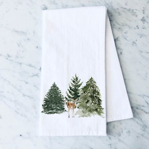 May include: A white kitchen towel with a watercolor illustration of three green pine trees and a brown deer in the center.