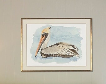 May include: A framed watercolor painting of a brown pelican with a light blue background. The pelican is resting on its belly with its head turned to the side. The painting is hanging on a wall above a light gray couch with two pillows.
