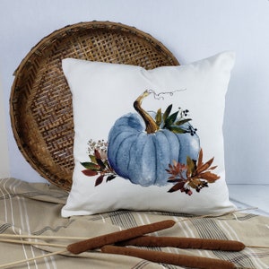 May include: A white pillow with a blue watercolor pumpkin and fall foliage design. The pillow is sitting on a woven basket and a striped fabric.