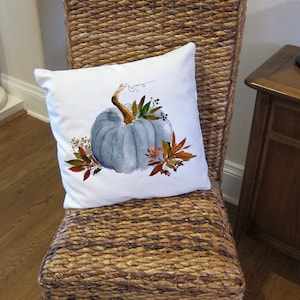 May include: A white throw pillow with a watercolor painting of a blue pumpkin with fall leaves and berries. The pillow is on a wicker chair with a brown wooden frame.