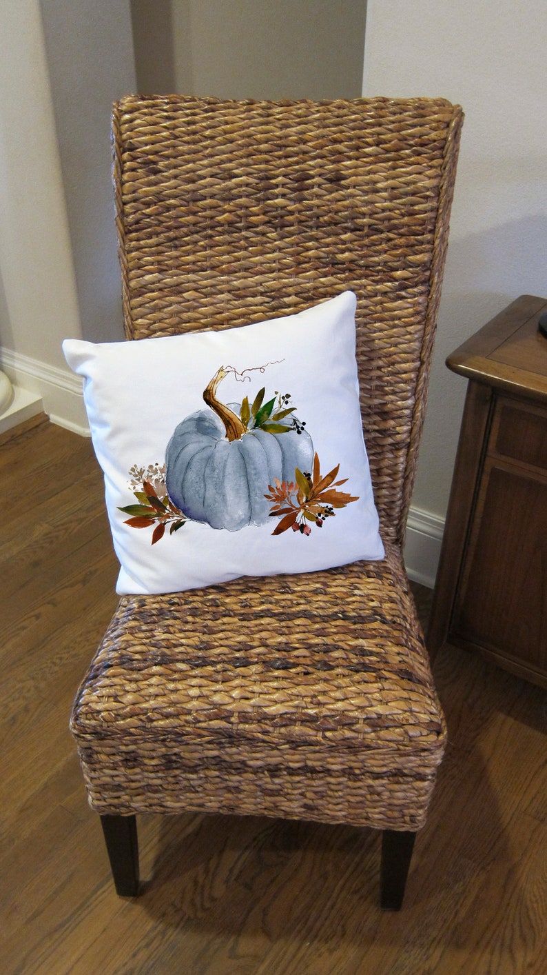 May include: A white throw pillow with a watercolor painting of a blue pumpkin with fall leaves and berries. The pillow is on a wicker chair with a brown wooden frame.