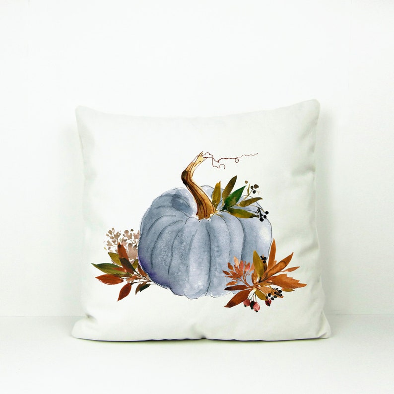 May include: A white pillow with a watercolor illustration of a blue pumpkin with brown stems and leaves. The pumpkin is surrounded by brown and green leaves and berries.