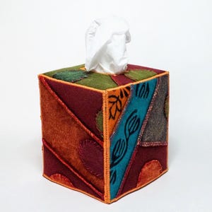 May include: A square tissue box cover with a patchwork design. The cover features various colors, including burgundy, orange, teal, and green, with decorative stitching. White tissues are visible at the top.