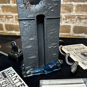 May include: A black 3D printed model of a broken tower with a blue water base. The tower is surrounded by a newspaper with the headline "Mass Breakout Azkaban" and a wanted poster for Newt Scamander.