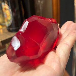 May include: A large, red, faceted, glass gemstone. The gemstone is smooth and shiny, with a translucent appearance.