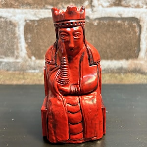 May include: A red chess piece depicting a queen wearing a crown and sitting on a throne. The piece is made of a smooth, glossy material and has a detailed design.