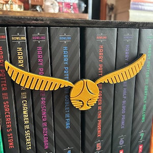 May include: A set of seven Harry Potter books with a golden snitch bookend. The books are arranged in order from left to right, with the first book, "Harry Potter and the Sorcerer's Stone", on the left and the seventh book, "Harry Potter and the Deathly Hallows", on the right.
