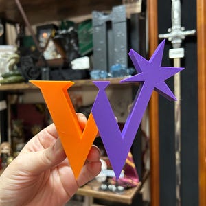 May include: A 3D printed letter W, with the left side orange and the right side purple. The letter is held in a hand.