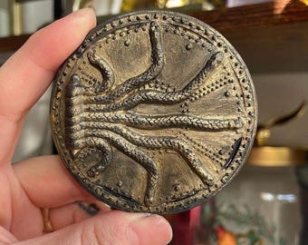 May include: A round, gold-colored metal medallion with a raised design of a snake with multiple heads. The medallion has a textured border.