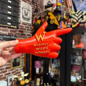 Puede incluir: Un artículo novedoso rojo brillante en forma de mano que señala, con el texto dorado "Weasley's Wizard Wheezes!". La letra "W" también es dorada. El artículo se sostiene frente a una exhibición de la tienda.