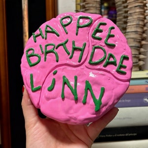 May include: Pink cake decorated with green icing. The cake is shaped like a brain and has the words "Happy Birthday Linn" written on it.