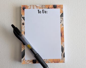 Può includere: Un blocco note rettangolare con un centro bianco e un bordo floreale con girasoli gialli e arancioni e foglie verde scuro. Il testo To Do: è stampato in nero nella parte superiore. Una penna gel nera retrattile poggia diagonalmente sul blocco.