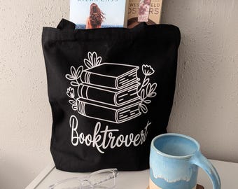 Può includere: Una borsa in tela nera con una grafica bianca di tre libri impilati circondati da disegni floreali a linee e il testo "Booktrovert" in corsivo. Accanto alla borsa ci sono occhiali con montatura trasparente e una tazza in ceramica blu e beige.