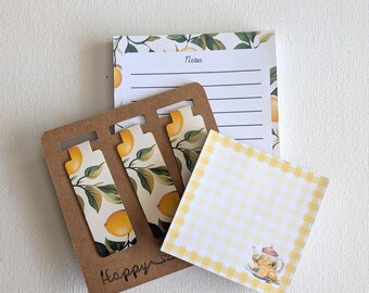 Può includere: Un set di cancelleria che include un blocco note a righe con bordo di limoni, tre segnalibri magnetici con illustrazioni di rami di limone su un supporto di cartone marrone con la parola Happy e un post-it a quadretti gialli con una teiera.