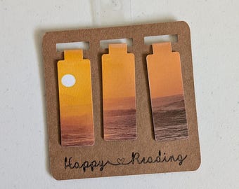Può includere: Tre segnalibri rettangolari con una scena di tramonto sull'oceano e un sole bianco a sinistra, fissati su un supporto di cartoncino marrone con il testo Happy Reading stampato in corsivo nero e un piccolo cuore tra le due parole.