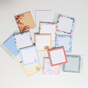 May include: A collection of square sticky note pads featuring various designs including floral patterns, gingham borders, and small illustrations of animals and objects. The pads are arranged in a scattered layout, with some encased in clear plastic sleeves.
