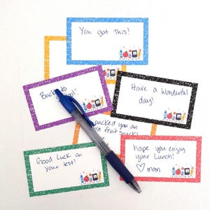 May include: A blue ballpoint pen resting on several rectangular lunchbox notes with colorful speckled borders. The notes feature handwritten messages like You got this, Back to school, Have a wonderful day, and Hope you enjoy your lunch, with small school supply illustrations.