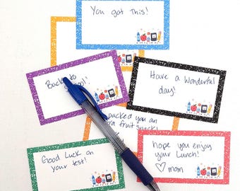 Può includere: Una penna a sfera blu appoggiata su diversi bigliettini rettangolari per il portapranzo con bordi colorati e punteggiati. I biglietti presentano messaggi scritti a mano come You got this, Back to school, Have a wonderful day e Hope you enjoy your lunch, con piccole illustrazioni di materiale scolastico.