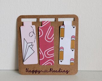 Peut inclure: Un support en carton brun contenant trois marque-pages rectangulaires. Le marque-page de gauche est rose clair avec un avion en papier, celui du centre est rose foncé avec des boucles blanches, et celui de droite est blanc avec des crayons jaunes. Le texte Happy Reading est imprimé en bas.