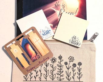 Può includere: Una pochette con cerniera in tela beige con stampa floreale nera e il testo ROOTED IN FAITH, un libro intitolato He is Risen, due blocchi note quadrati con testo religioso e un set di segnalibri in cartone marrone con tre croci.