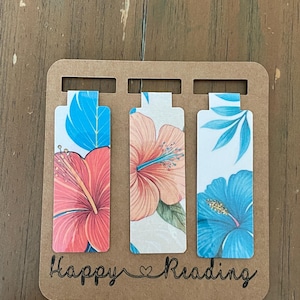 May include: A set of three floral bookmarks with a brown cardboard backing. Each bookmark features a different hibiscus flower design in shades of pink, orange, and blue. The words "Happy Reading" are written in cursive.