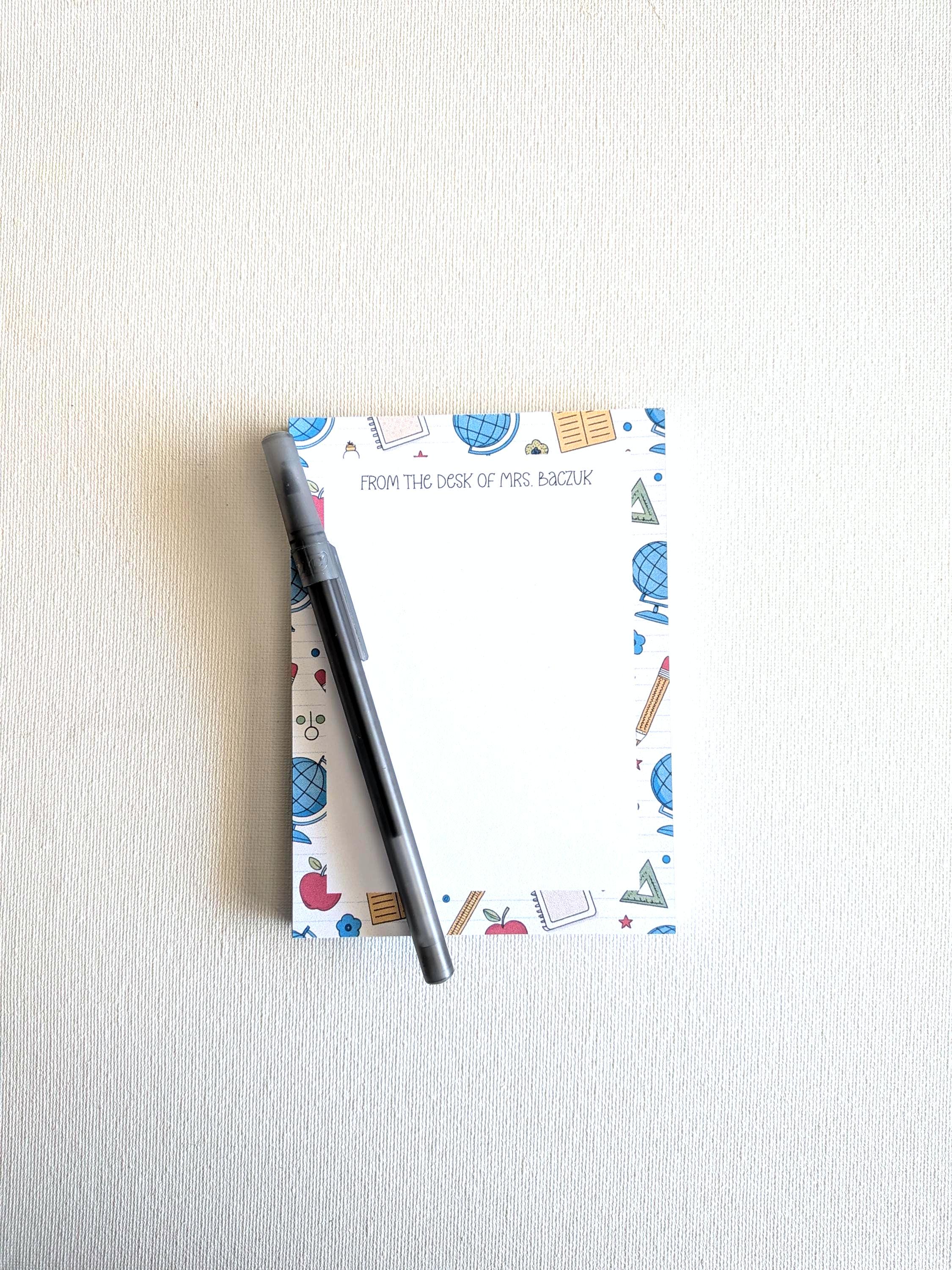 Può includere: Un blocco note rettangolare con un centro bianco e un bordo decorato con illustrazioni scolastiche colorate come mappamondi, mele e righelli. Il testo "FROM THE DESK OF MRS. BACZUK" è stampato in alto. Una penna grigio scuro è appoggiata in diagonale.