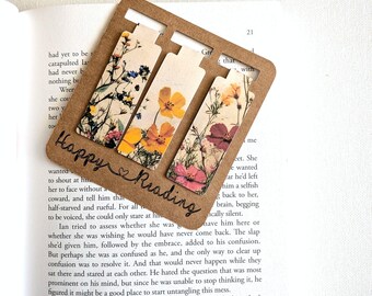 Può includere: Un supporto in cartone marrone contenente tre segnalibri rettangolari, ognuno caratterizzato da una stampa floreale colorata con fiori di campo nelle tonalità del giallo, rosa e blu. Il supporto riporta la scritta Happy Reading in corsivo nero.