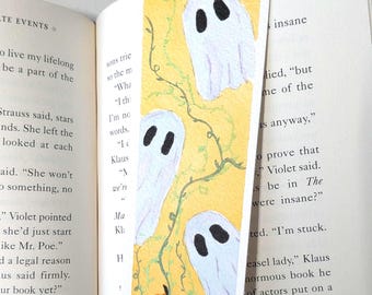 Può includere: Un segnalibro rettangolare in carta con sfondo giallo, tre fantasmi bianchi, viticci verdi intrecciati e due zucche arancioni sul fondo. L'illustrazione è dipinta in stile acquerello con occhi neri su ogni fantasma.