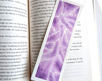 Può includere: Un segnalibro rettangolare in carta con un dipinto ad acquerello di forme astratte viola e lavanda separate da sottili linee bianche, appoggiato su un libro aperto con testo stampato e il numero di pagina 143 visibile in basso a destra.