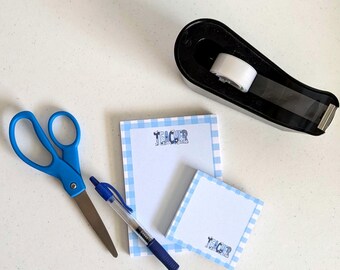 Peut inclure: Un ensemble de papeterie comprenant des ciseaux bleus, un stylo à bille bleu, un dévidoir de ruban adhésif noir et deux blocs-notes blancs à bordure vichy bleu clair. Le mot TEACHER est imprimé en bleu sur les deux blocs.