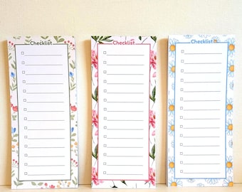Può includere: Tre blocchi note rettangolari con bordi floreali. Il blocco a sinistra ha un bordo verde con piccoli fiori, quello centrale un bordo rosa con peonie e quello a destra un bordo blu con margherite. Ognuno riporta la parola Checklist.
