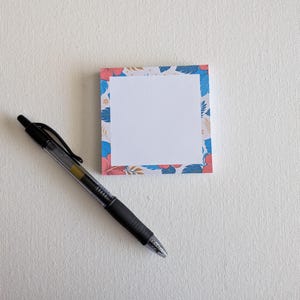 May include: A square sticky note pad featuring a white center and a border decorated with blue and pink tropical hibiscus flowers and green leaves, accompanied by a black retractable gel ink pen positioned diagonally on the left side.