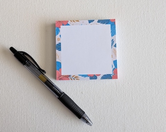 Può includere: Un blocco di foglietti adesivi quadrati con un centro bianco e un bordo decorato con fiori di ibisco tropicali blu e rosa e foglie verdi, accompagnato da una penna a inchiostro gel nera retrattile posizionata in diagonale a sinistra.