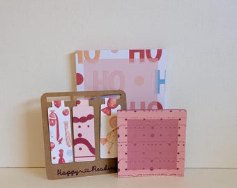 Può includere: Un set di cancelleria che include un blocco note quadrato con il testo HO HO, un supporto in cartone marrone contenente tre segnalibri verticali con illustrazioni di caramelle e un blocco note rosa quadrato con un motivo a pois e smerlato.